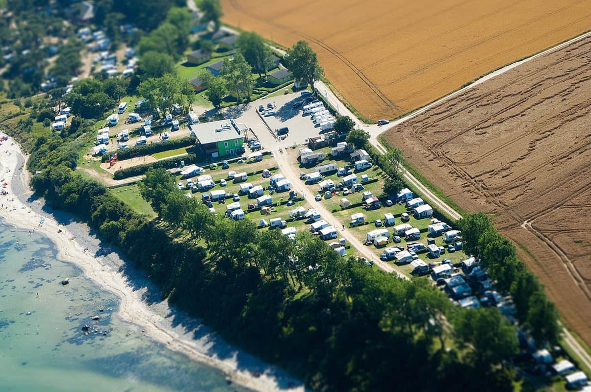 Luftbildaufnahme Luftaufnahme eines Küstencampingplatzes mit zahlreichen Zelten und Wohnwagen, gesäumt von Bäumen und Feldern, neben einem Sandstrand und klarem, blauem Wasser.