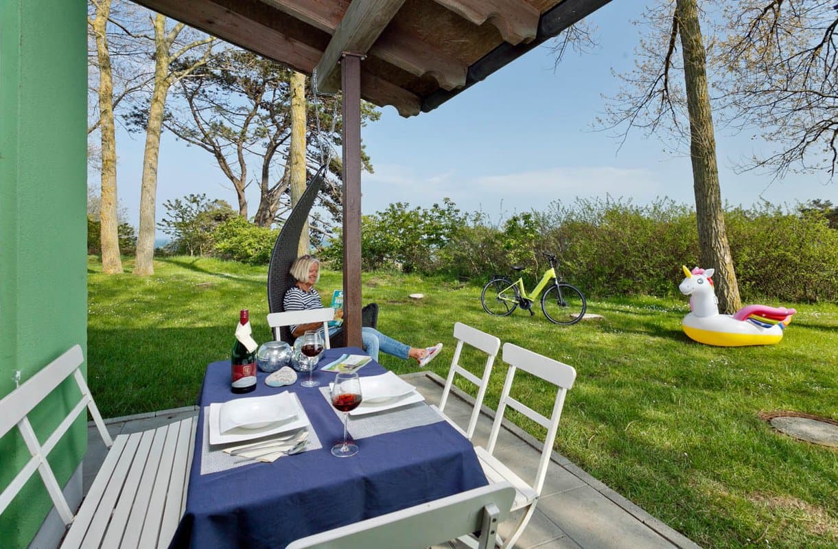 Ferienhäuser Terrasse Eine Person entspannt sich in einer Hängematte in einem Garten, mit einem gedeckten Tisch, einem Fahrrad und einem aufblasbaren Einhorn im Hintergrund.