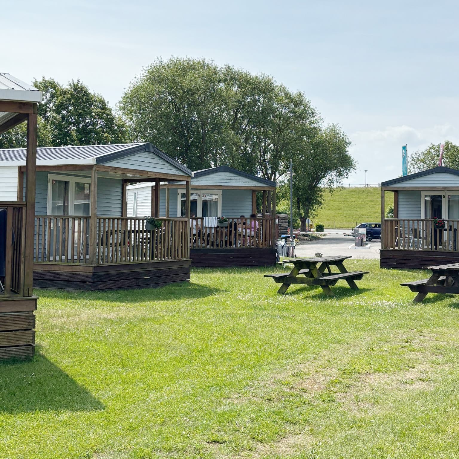 Kleine Hütten mit Holzveranden auf einer Wiese, umgeben von Picknicktischen und Bäumen unter einem klaren Himmel.