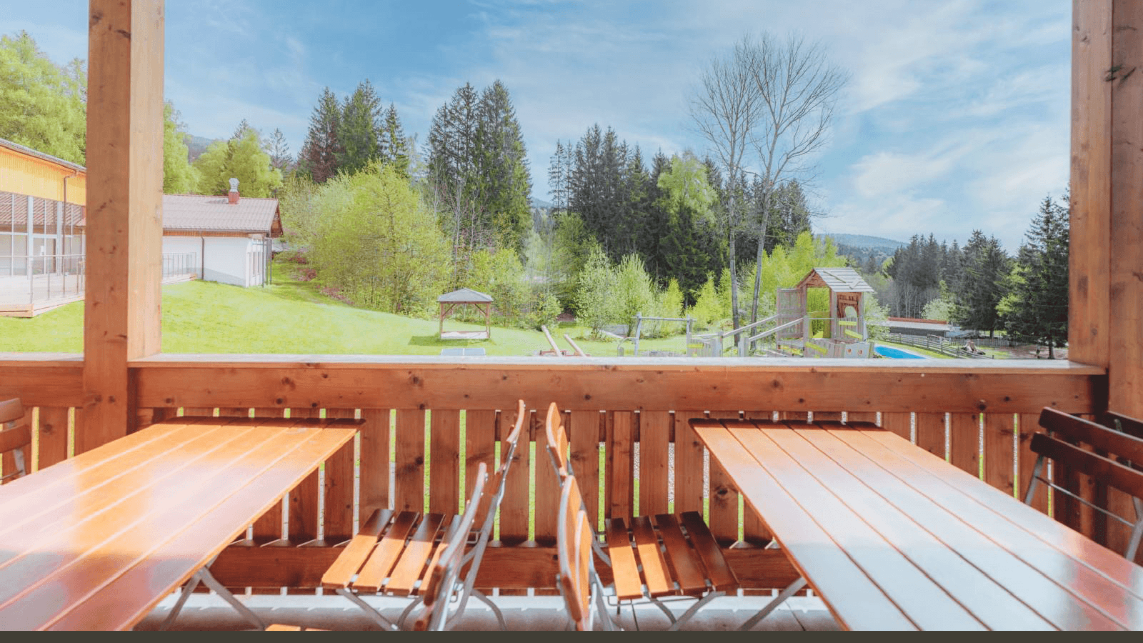 Restaurant Holztische und -stühle auf einer Terrasse mit Blick auf eine üppige grüne Landschaft mit Bäumen, einem Spielplatz und einem klaren blauen Himmel im Hintergrund.