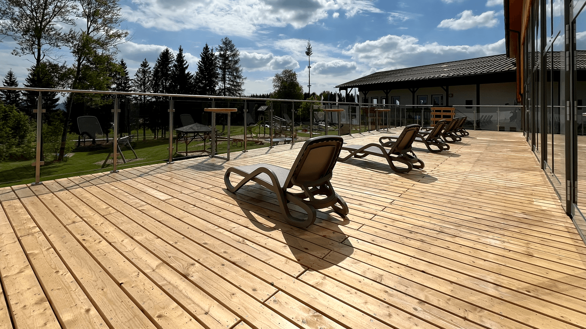 Sonnenterasse Holzdeck mit mehreren Liegestühlen mit Blick auf Bäume und einen teilweise bewölkten Himmel, angrenzend an ein modernes Gebäude.