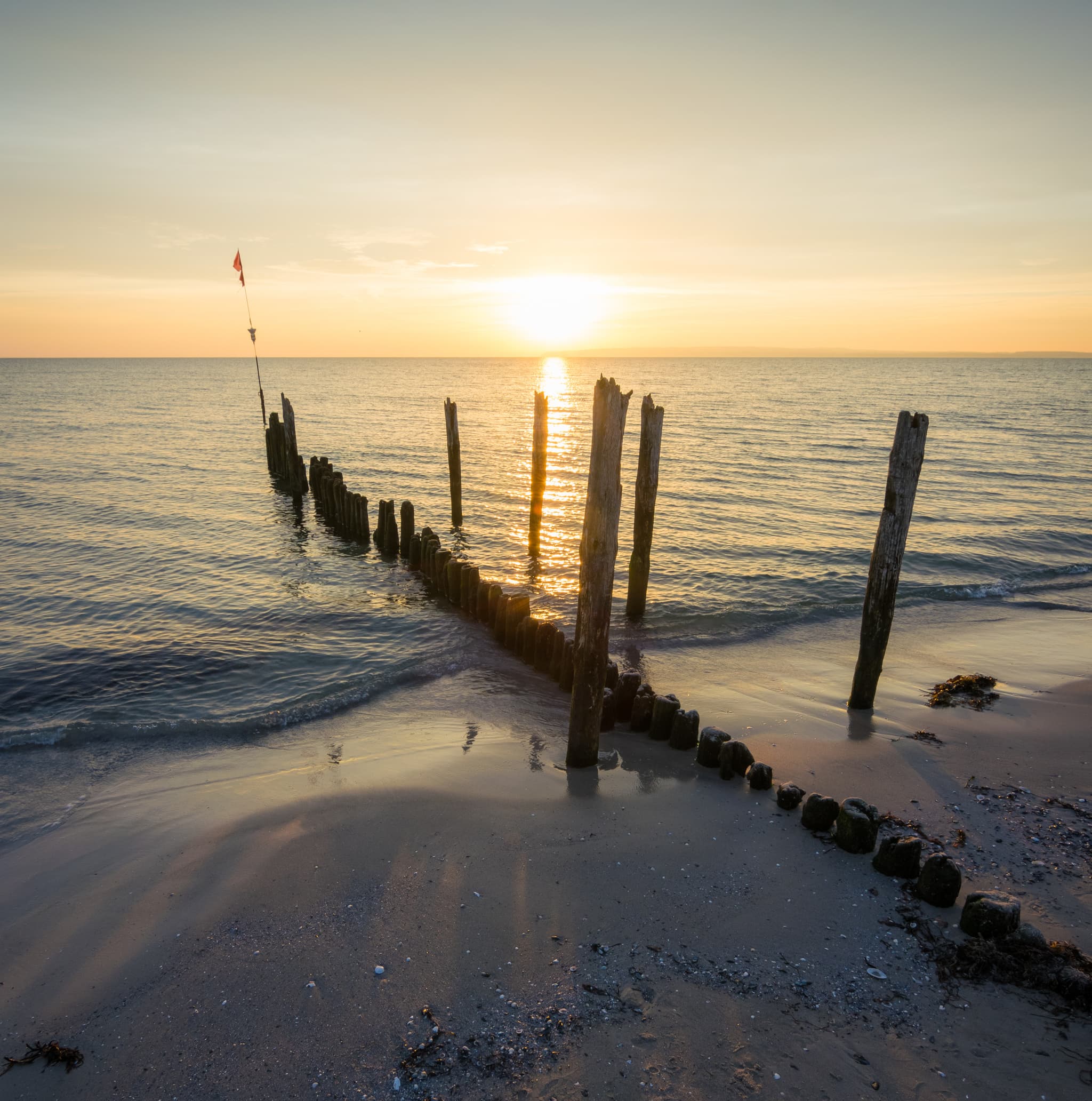Rügen Sonnenuntergang über einer ruhigen See mit verwitterten Holzpfählen, die von einem Sandstrand ins Wasser ragen und lange Schatten werfen.