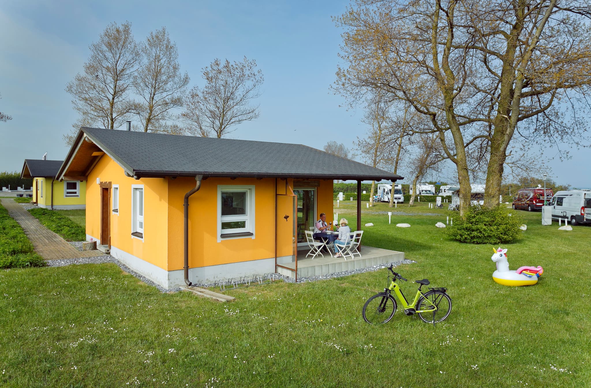 A small orange house with a porch, surrounded by grass and trees. A bicycle and inflatable unicorn are on the lawn. People sit at a table.