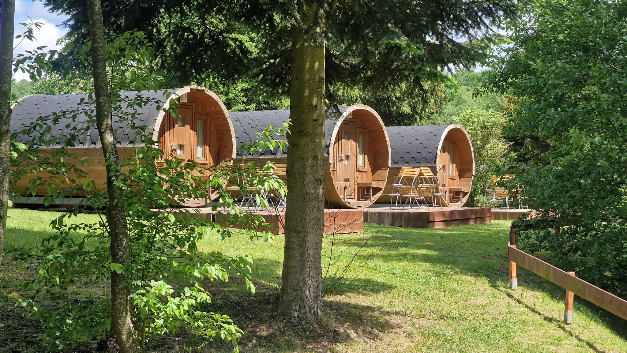Drei fassförmige Holzhütten auf einer von Bäumen umgebenen Wiese mit Sitzgelegenheiten auf den Decks.