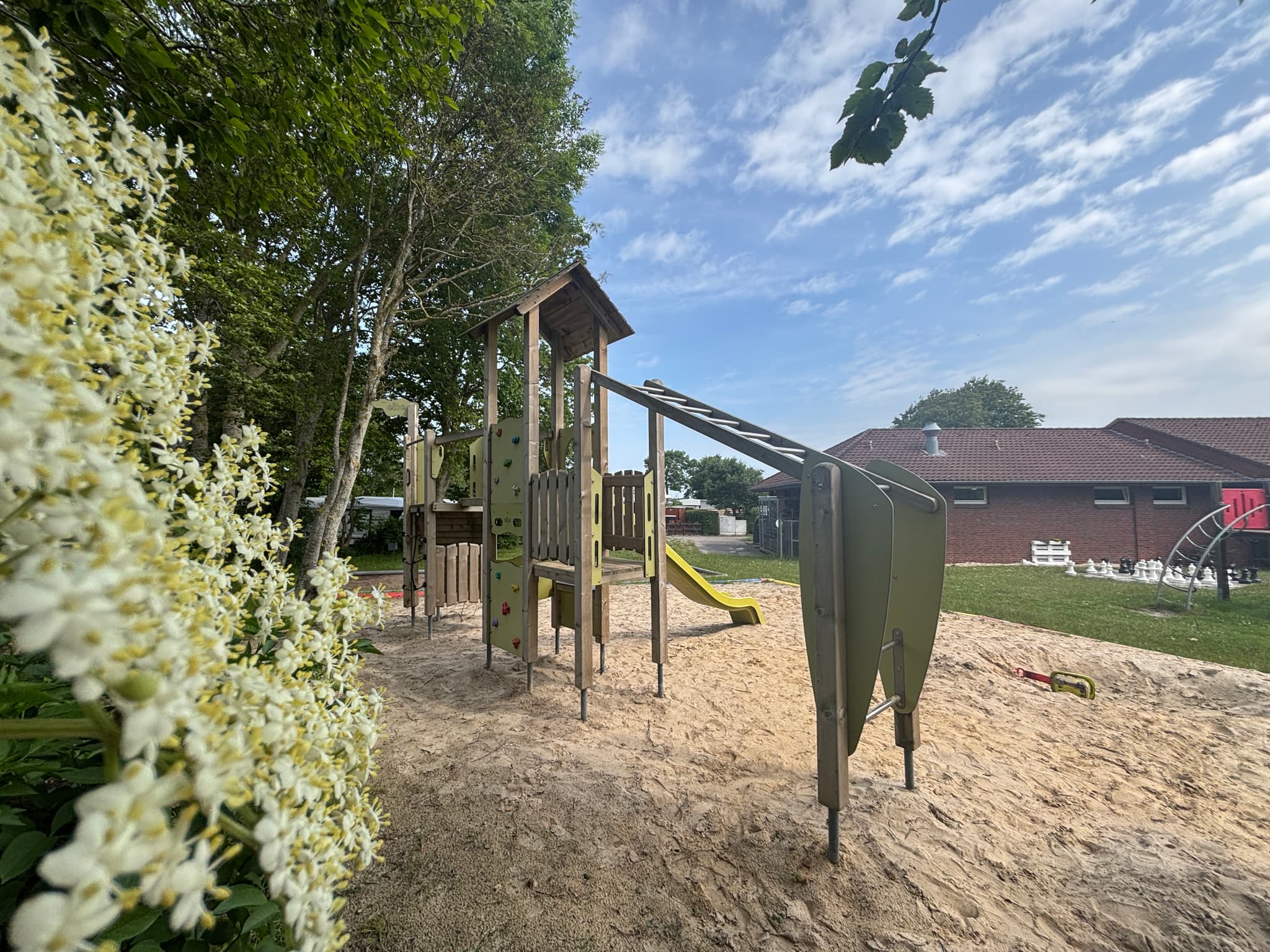 Spielplatz Spielplatz mit einem Klettergerüst und einer Rutsche auf sandigem Boden, umgeben von Bäumen und einem Gebäude im Hintergrund unter einem teilweise bewölkten Himmel.
