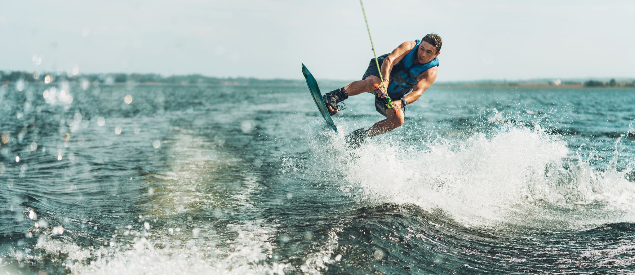 Wakeboard Eine Person in einer blauen Weste springt beim Wakeboarden auf einem See, umgeben von plätscherndem Wasser und unter einem klaren Himmel.