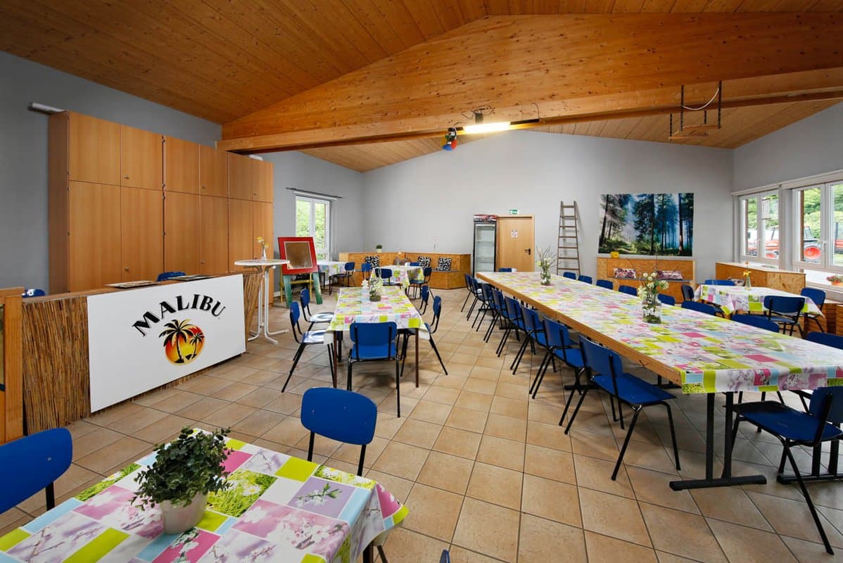 Aufenthaltsraum Gemeindesaal mit bunten Tischtüchern, blauen Stühlen, einem „Malibu“-Schild und Topfpflanzen. Große Fenster und eine Holzdecke schaffen eine helle Atmosphäre.