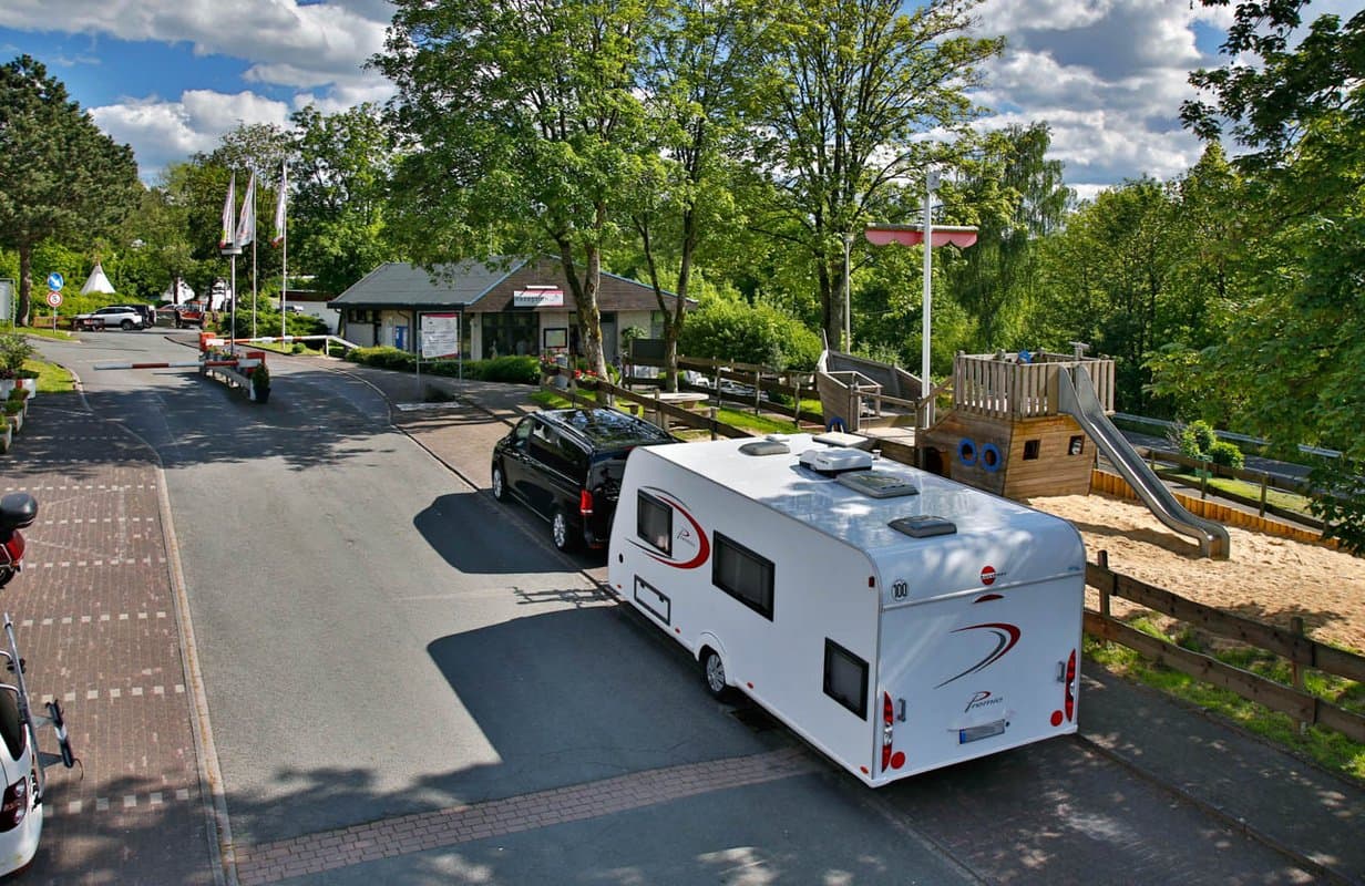 Anfahrt Rezeption Auto, das einen weißen Wohnwagen zieht, auf einer Straße in der Nähe eines Spielplatzes mit einer Rutsche, umgeben von Bäumen und Gebäuden unter einem teilweise bewölkten Himmel.