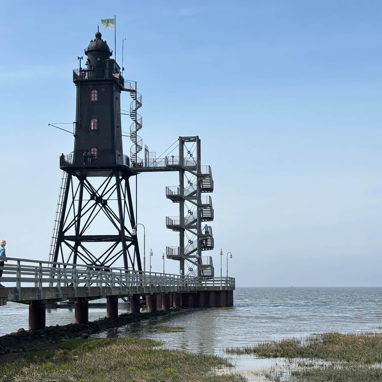 Leuchtturm Ein hoher, schwarzer Leuchtturm auf Stelzen mit einer Wendeltreppe, verbunden mit einem Steg über ruhigem Wasser unter einem klaren blauen Himmel.
