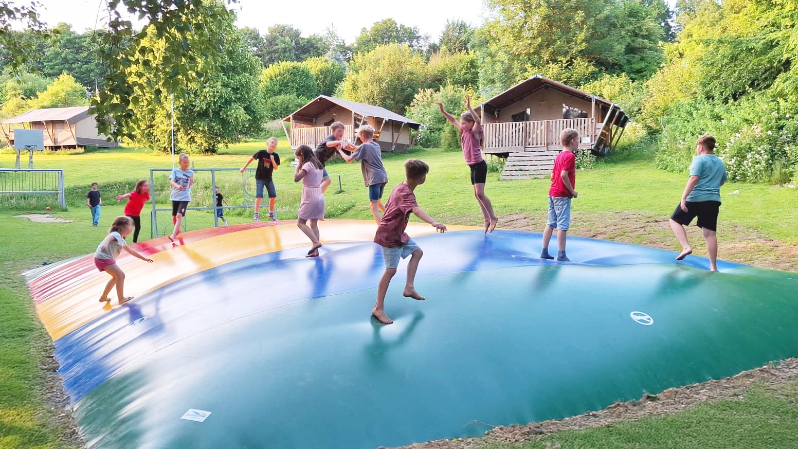 Kinder spielen und springen auf einer bunten aufblasbaren Hüpfburg auf einer Wiese mit Bäumen und Hütten im Hintergrund.
