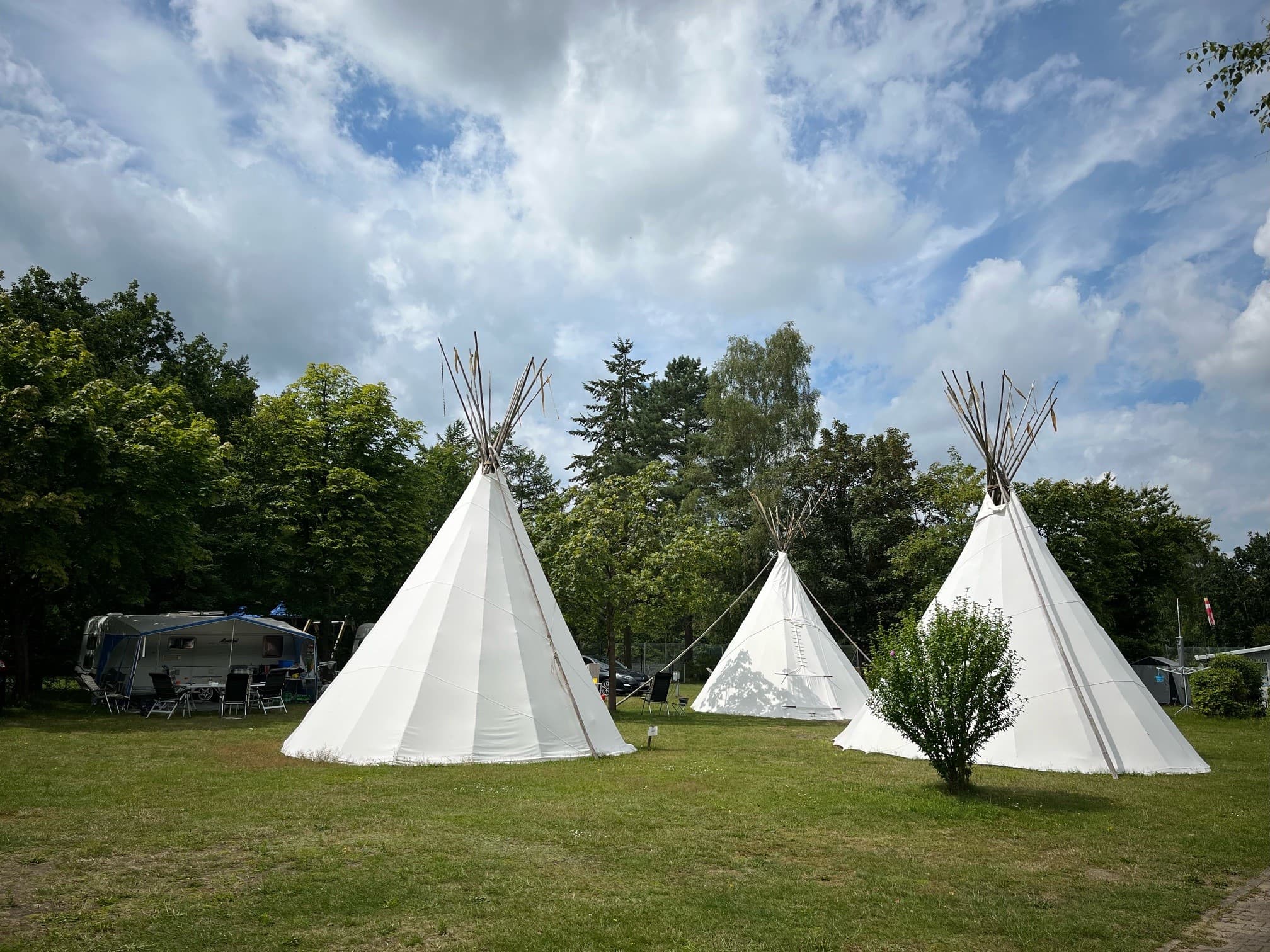 Tipi Drei weiße Tipis auf einer von Bäumen umgebenen Wiese unter einem teilweise bewölkten Himmel, mit einem Wohnwagen und Stühlen in der Nähe.