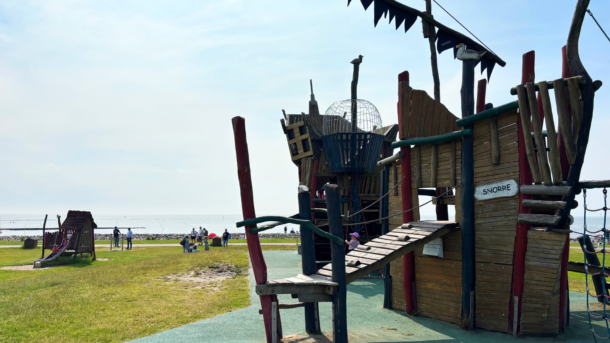 Spielplatz Spielplatz mit einem hölzernen Piratenschiff Struktur auf Gras in der Nähe eines Strandes, mit Menschen im Hintergrund unter einem bewölkten Himmel.