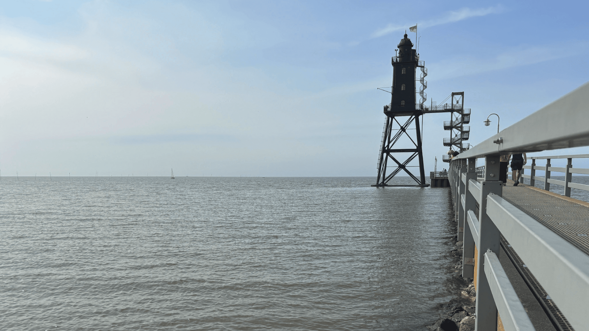 Ein Leuchtturm steht auf einer Metallkonstruktion über ruhigem Wasser, zu dem ein Geländer und ein Steg unter klarem Himmel führen.