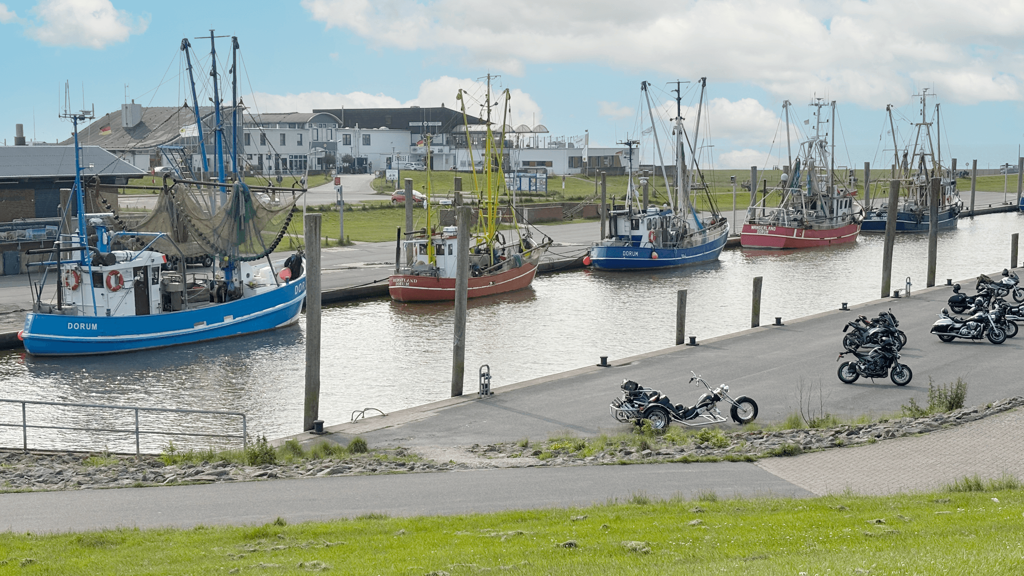 An einem sonnigen Tag mit teilweise bewölktem Himmel liegen Fischerboote in einem ruhigen Hafen vor Anker, in dessen Nähe Motorräder geparkt sind.