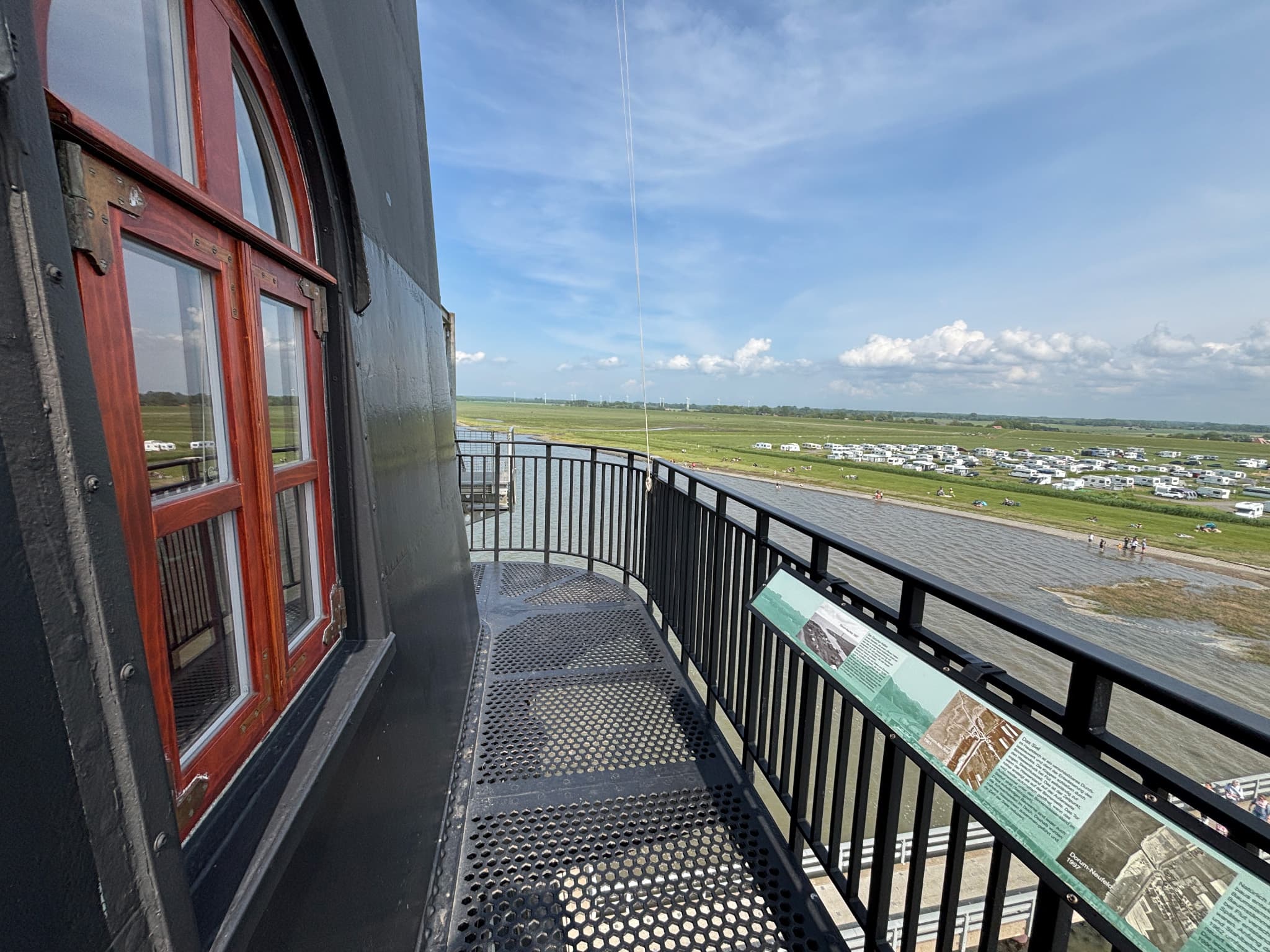 Blick vom Balkon eines Leuchtturms auf eine Graslandschaft und einen Strand mit geparkten Wohnmobilen unter einem blauen Himmel mit vereinzelten Wolken.
