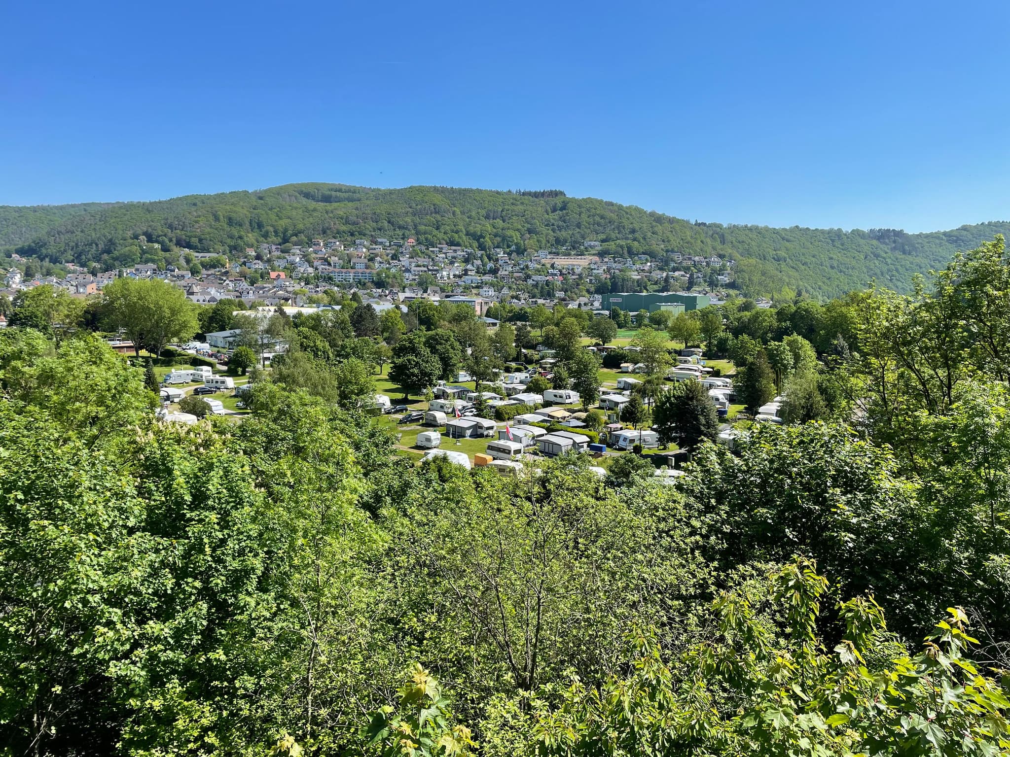 Ein malerischer Blick auf ein üppig grünes Tal mit zahlreichen weißen Wohnwagen und einer Stadt, die sich am Fuße eines bewaldeten Hügels unter einem klaren blauen Himmel befindet.