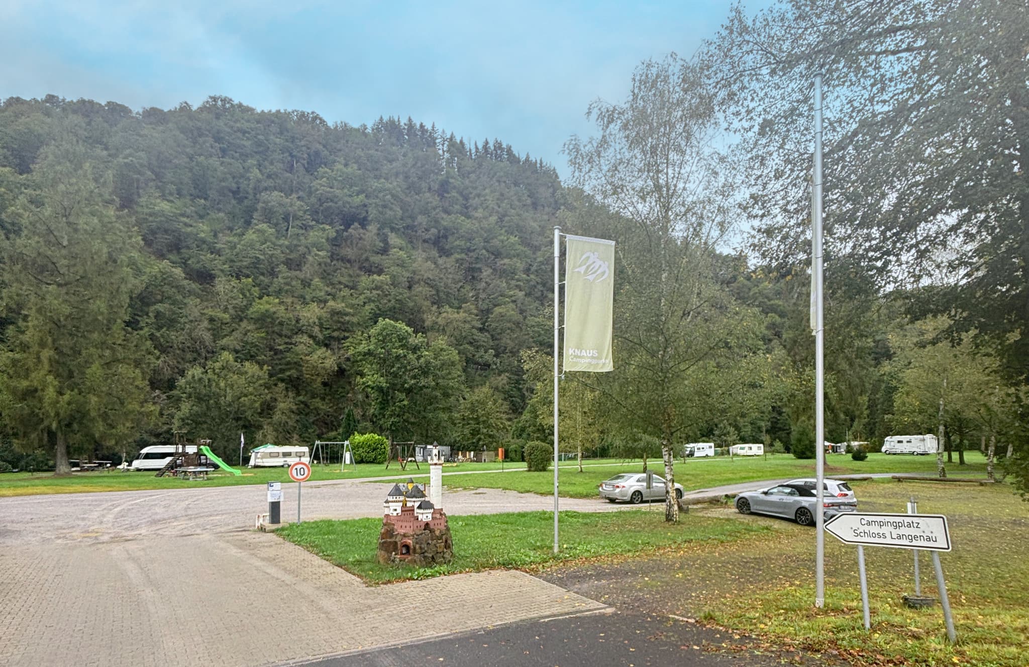 Einfahrt Campingplatz mit geparkten Wohnwagen, Autos und einem Spielplatz. Umgeben von üppigen grünen Bäumen und Hügeln unter einem bewölkten Himmel. Schilder am Eingang sichtbar.
