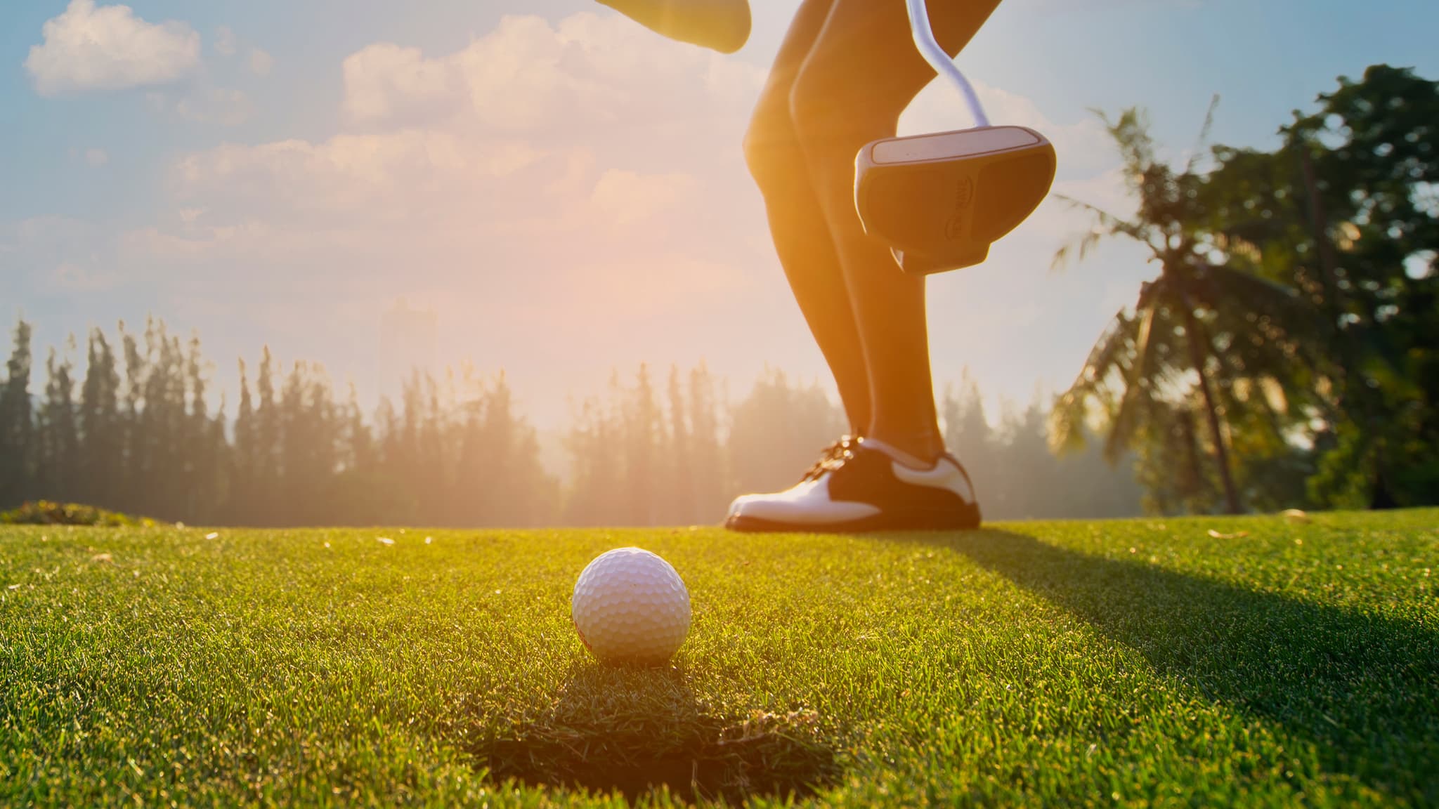Golfer, der sich auf einem sonnigen Platz auf den Putt vorbereitet, mit einem Golfball in der Nähe des Lochs und Bäumen im Hintergrund.