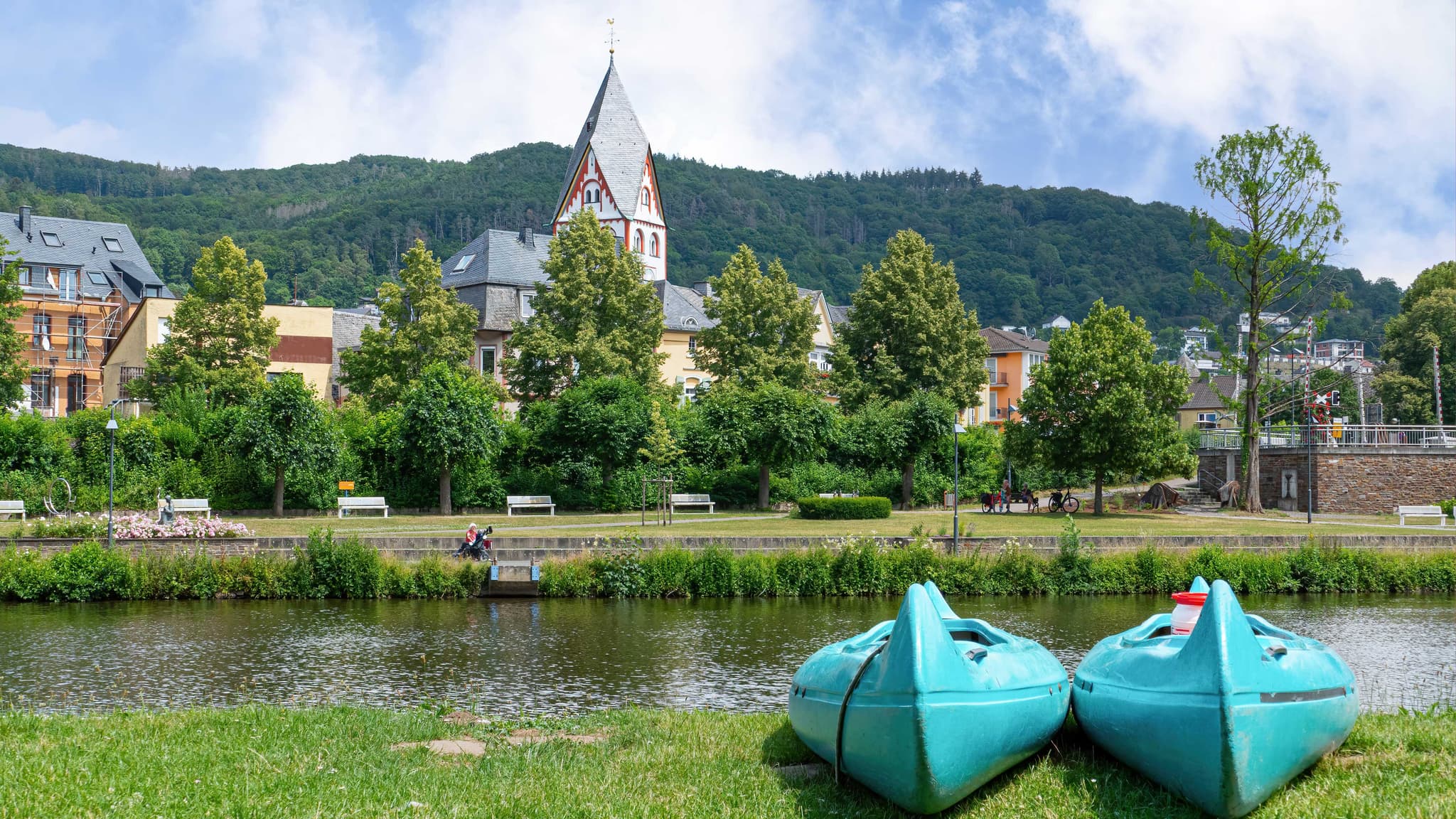 Nassau Zwei türkisfarbene Kajaks an einem grasbewachsenen Ufer eines Flusses, mit Blick auf eine Stadt, Bäume und einen Kirchturm im Hintergrund.