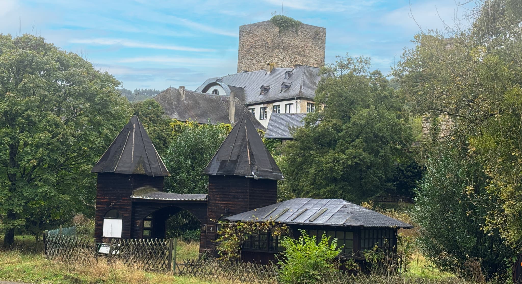 Schloss Eine rustikale Holzkonstruktion mit spitzen Dächern in einer üppigen Landschaft, mit einem Steinturm und einem Gebäude, die teilweise im Hintergrund sichtbar sind.
