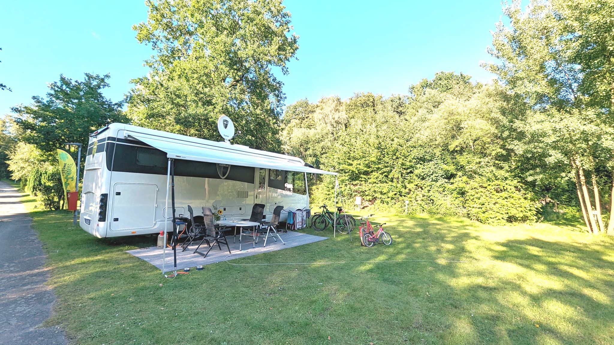 XXL Stellplätze Ein geparktes Wohnmobil mit ausgefahrener Markise, umgeben von Stühlen, einem Tisch und Fahrrädern auf einer Wiese mit Bäumen im Hintergrund.