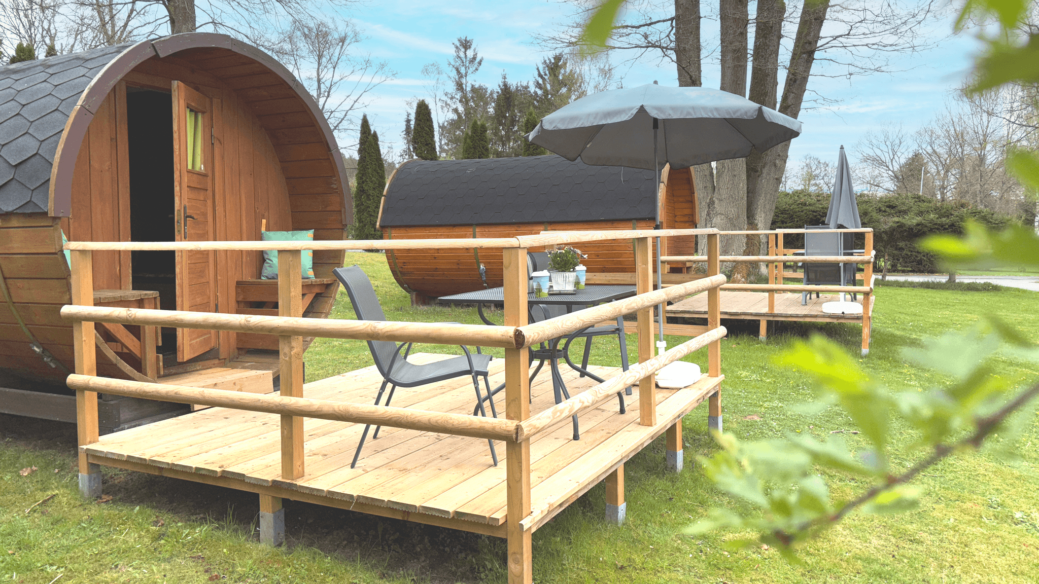 Holzhütten in Fassform mit kleinen Terrassen, Stühlen, Tischen und Sonnenschirmen auf einer Wiese mit Bäumen.