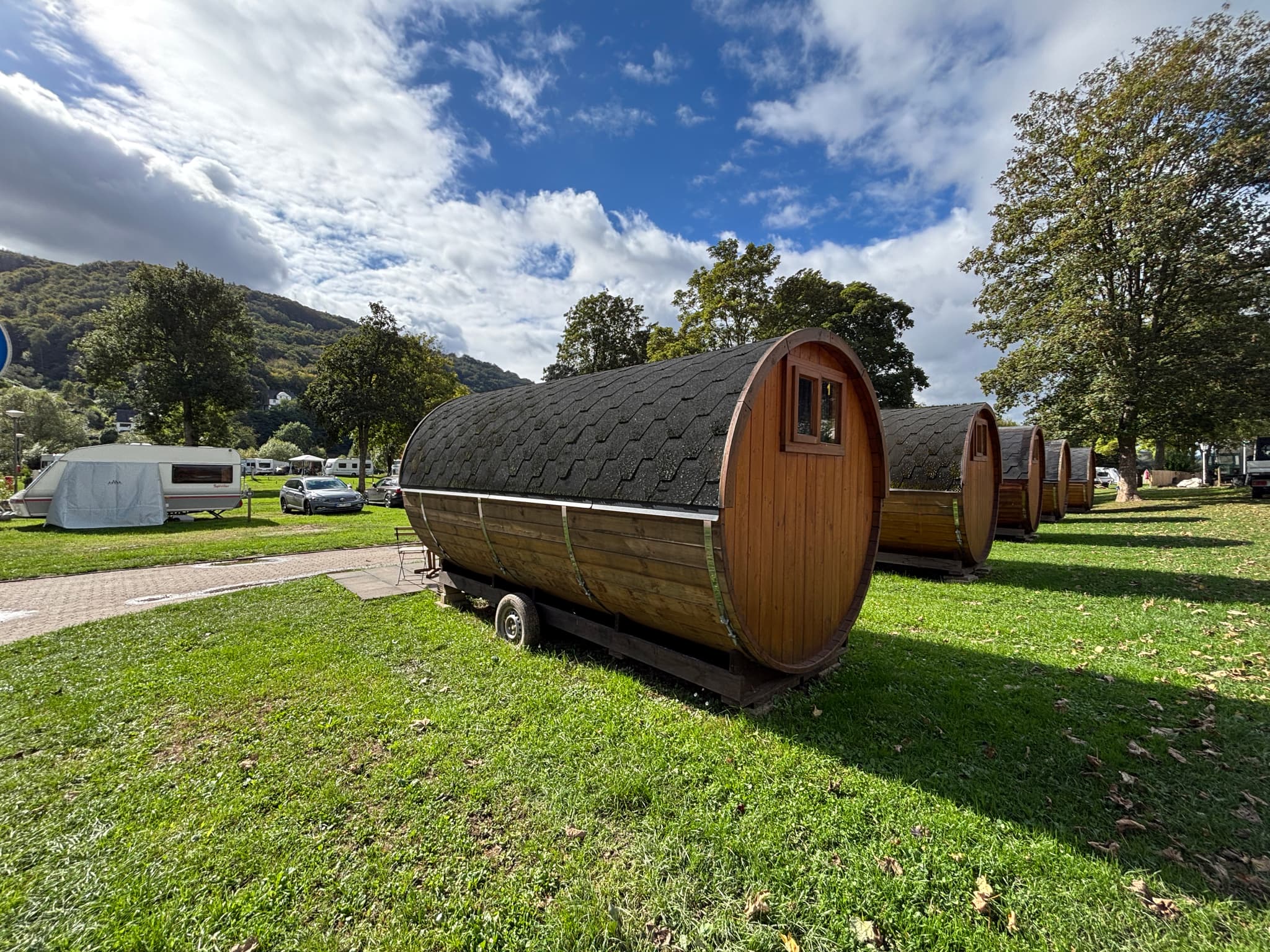 Fässer Faßförmige Holzhütten mit Schindeldächern auf einem grasbewachsenen Campingplatz, umgeben von Bäumen und Hügeln unter einem teilweise bewölkten Himmel.
