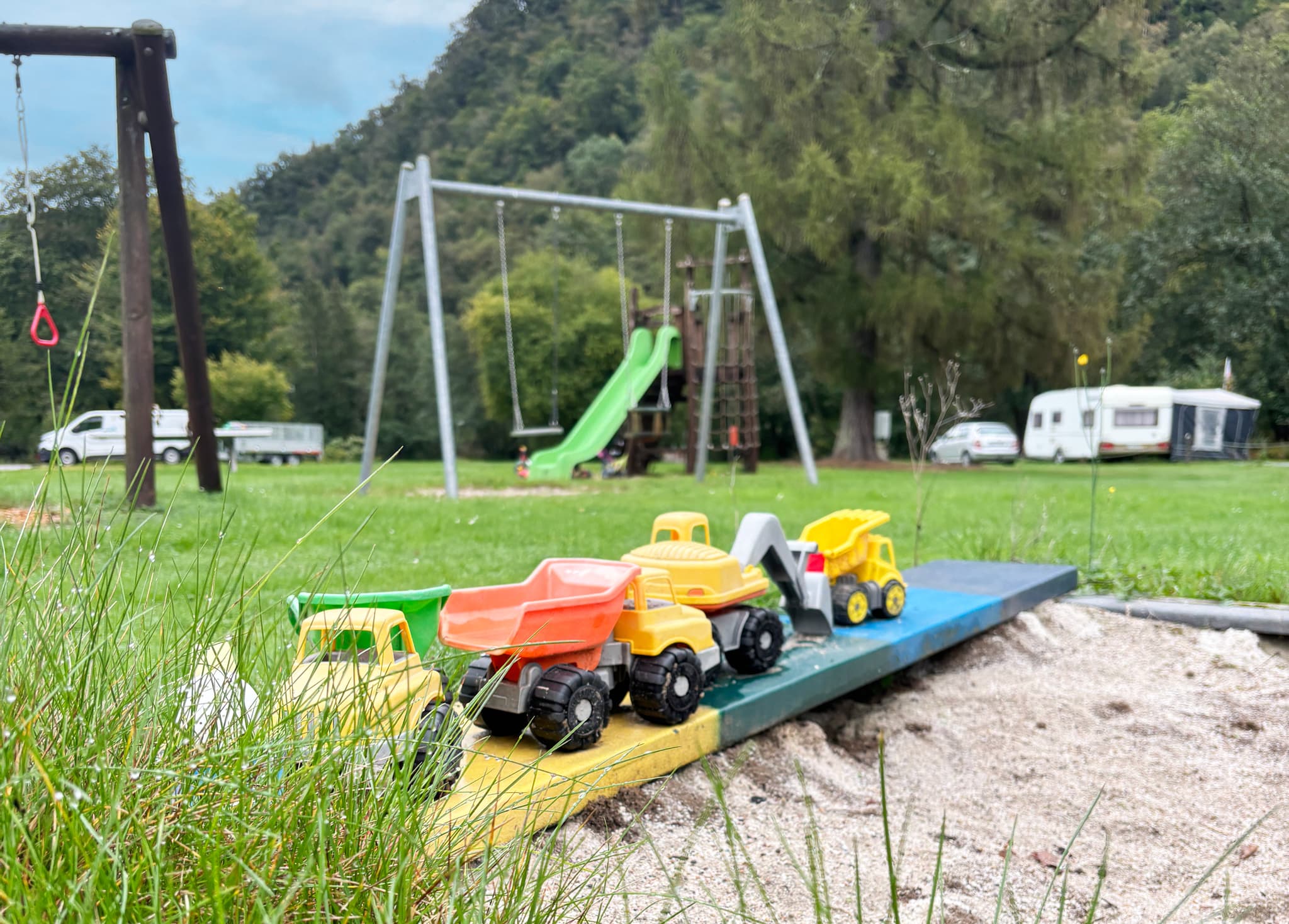 Spielplatz Spielzeugautos auf einer Sandkastenbrücke auf einem Spielplatz mit einer Schaukel, einer Rutsche, einer Wiese und geparkten Wohnwagen im Hintergrund.