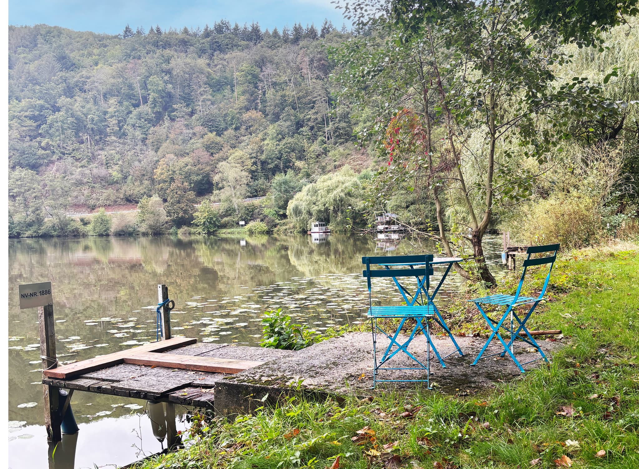 Parkromantik Zwei blaue Stühle und ein Tisch auf einer Terrasse am See, umgeben von Bäumen und Grün, mit einem kleinen Steg, der ins Wasser hineinragt.