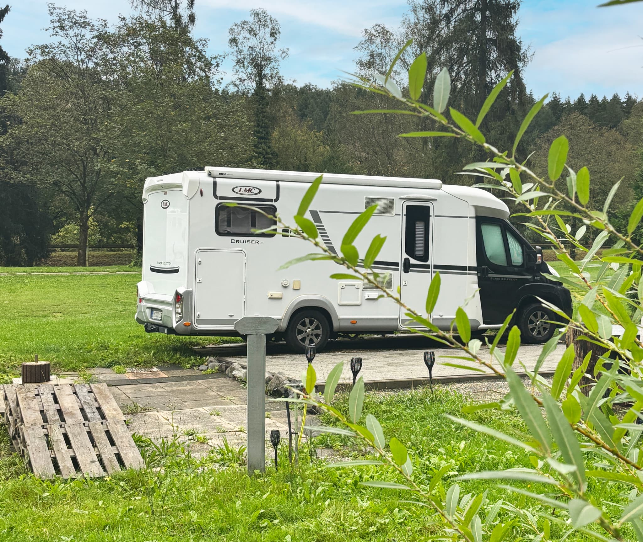 Stellplatz Ein weißes Wohnmobil, geparkt auf einer Wiese mit Bäumen im Hintergrund, umgeben von üppigem Grün und einem klaren blauen Himmel.