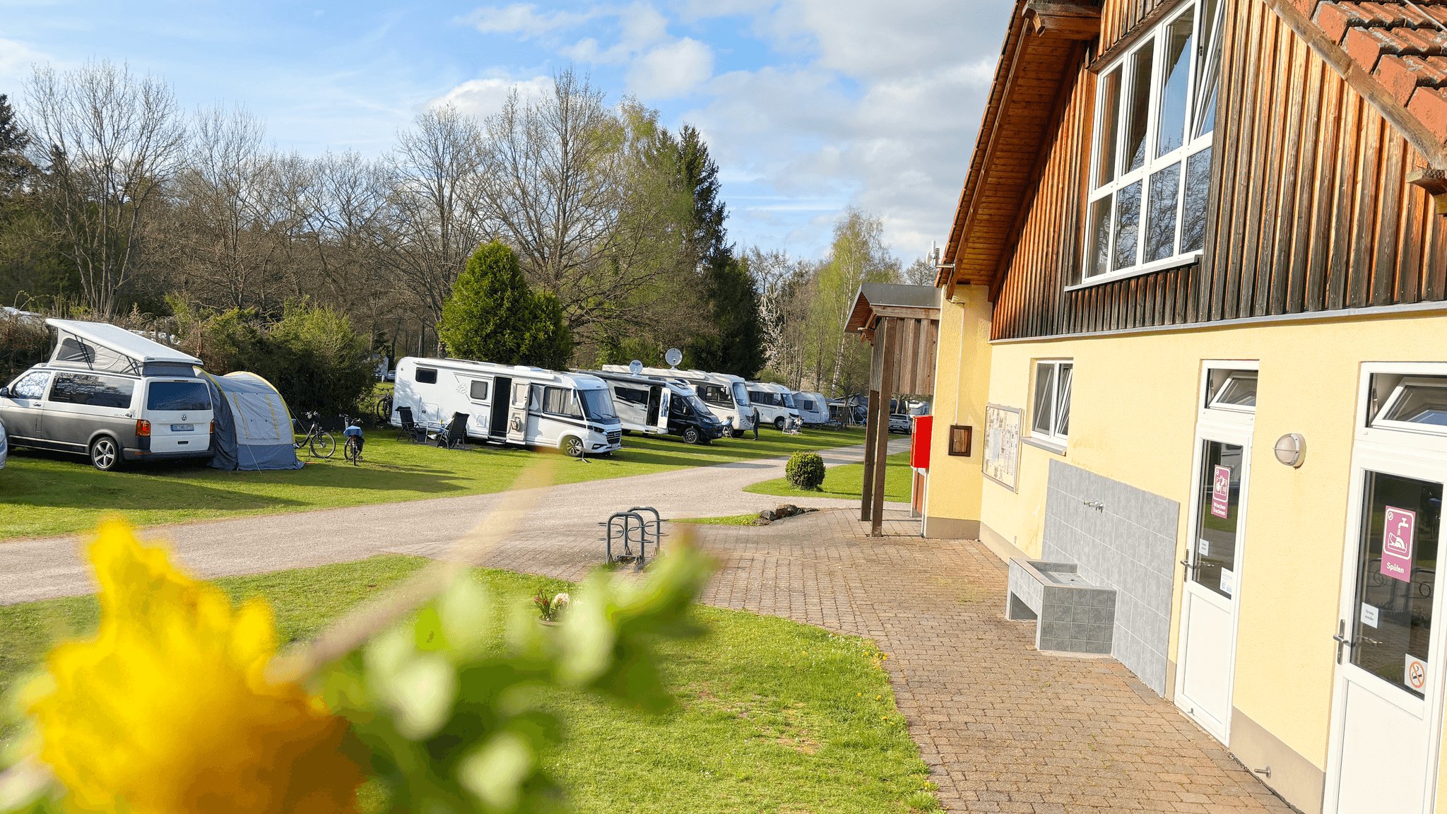 Ein Campingplatz mit mehreren geparkten Wohnmobilen und einem Zelt, daneben ein Holzgebäude. Im Vordergrund ist eine gelbe Blume zu sehen.