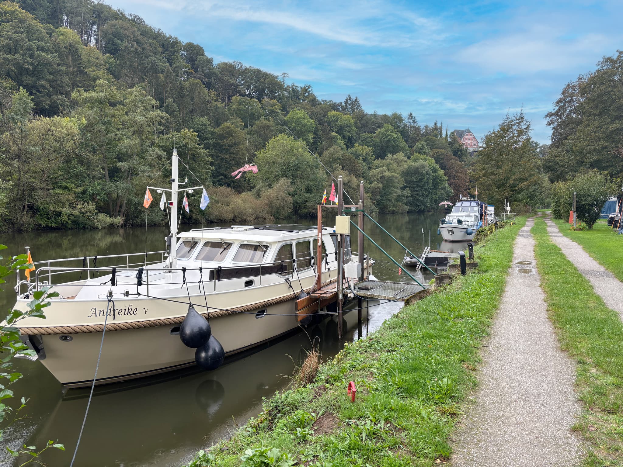 Uferweg Boote, die an einem ruhigen Fluss mit einem grasbewachsenen Weg und einer bewaldeten Fläche angedockt sind, unter einem teilweise bewölkten Himmel.