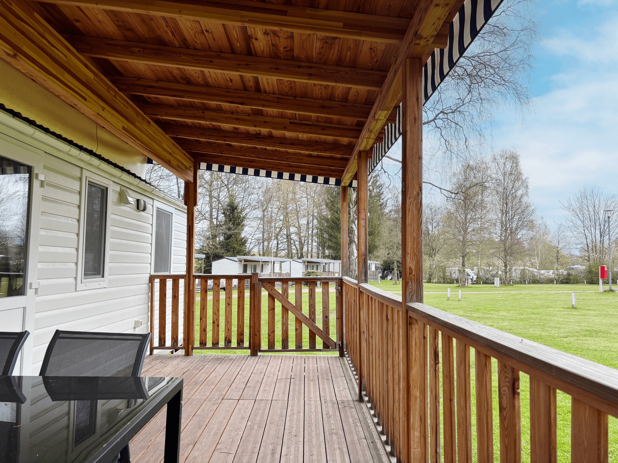 Hölzerne Veranda mit gestreifter Markise an einem weißen Wohnmobil, mit Blick auf eine Grasfläche mit Bäumen und anderen Wohnmobilen in der Ferne.