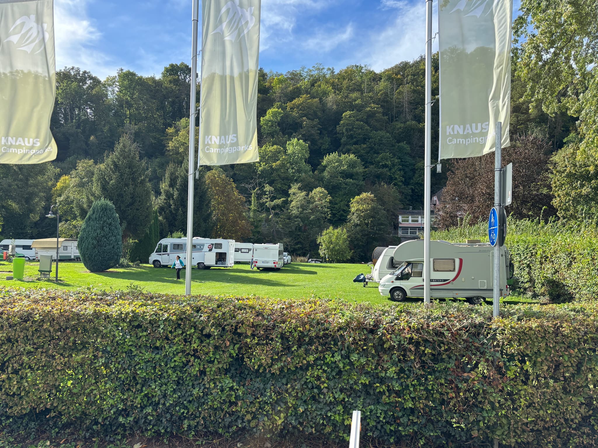 Stellplätze Campingplatz mit mehreren Wohnwagen auf einer Wiese, umgeben von Bäumen und Hügeln unter blauem Himmel, mit hohen Fahnen im Vordergrund.