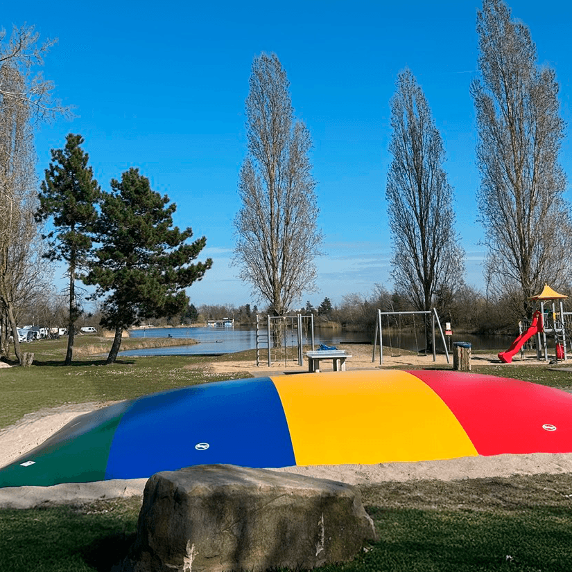 Hüpfkissen Colorful inflatable play structure in a park with tall trees, a swing set, and a slide. A lake is visible in the background under a clear blue sky.