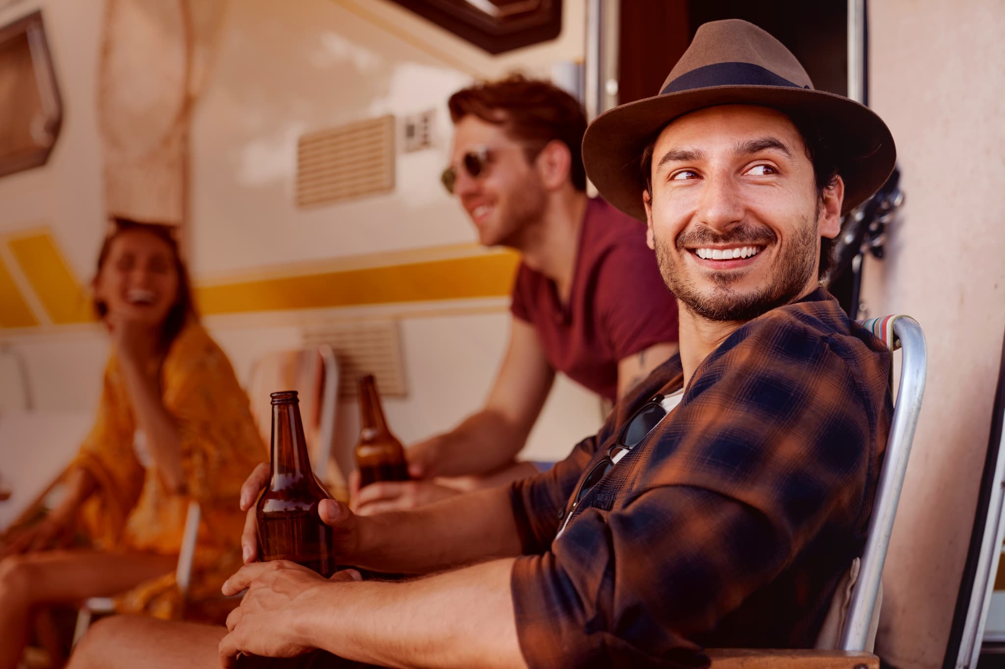 Freunde Drei Freunde sitzen vor einem Wohnmobil, lächeln und halten Bierflaschen in der Hand. Ein Mann mit einem Hut steht im Vordergrund und schaut zur Seite.