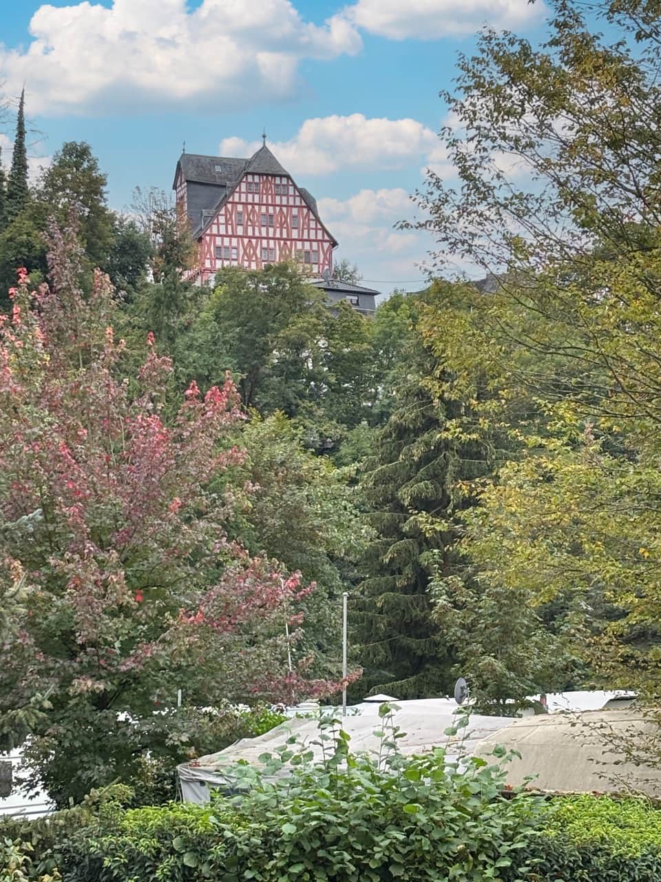 Nassau Ausblick Ein traditionelles Fachwerkhaus mit steilem Dach ist teilweise durch üppiges grünes und rotes Laub unter einem blauen Wolkenhimmel sichtbar.